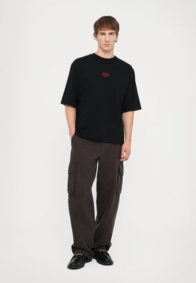 T-shirt noir surdimensionné avec un texte rouge, associé à un pantalon cargo ample marron foncé avec des poches latérales et une texture lisse.