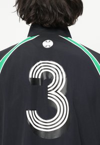 Person med kort mørkt hår iført en svart jakke med grønne striper og et stort hvitt nummer 3 på ryggen.