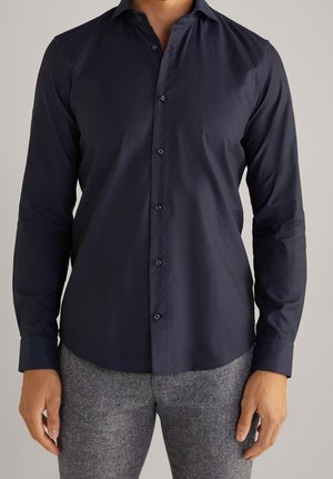 Mężczyzna ubrany w dopasowaną, granatową koszulę na guziki z długim rękawem oraz szare, fakturowane spodnie, stojący na jednolitym tle.