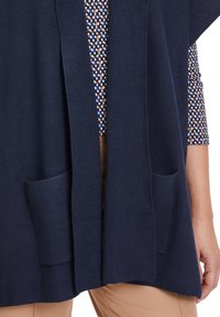 Marinblå stickad cardigan med öppen framsida, två framfickor och ribbad struktur. Bärs över en mönstrad skjorta i blå, brun och vit färg.