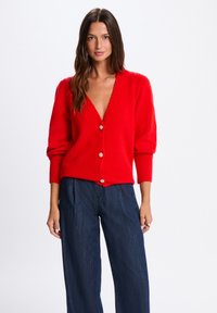 Roter Strickcardigan mit tiefem V-Ausschnitt, langen Ärmeln und gerippten Bündchen. Verfügt über silberne Knöpfe und einen lockeren Schnitt, kombiniert mit dunklen Jeans.