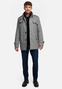 Grå uldblandet frakke med høj krave, knaplukning foran og sidelommer. Båret over en sort sweater og kombineret med mørke jeans og sorte støvler.