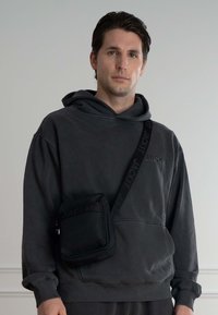 Sort crossbody taske med tekstureret stof og præget logo, båret over en mørkegrå hættetrøje med afslappet pasform.