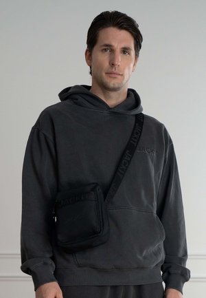 Zwarte crossover tas met gestructureerde stof en reliëflogo, gedragen over een donkergrijze, capuchontrui met een relaxte pasvorm.