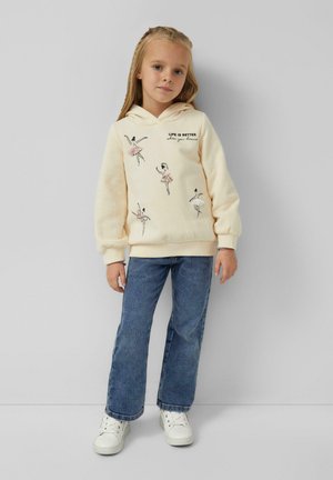 Junges Mädchen trägt einen cremefarbenen Hoodie mit Balletttänzerinnen-Illustrationen und Text, blaue Jeans und weiße Turnschuhe und steht vor einer schlichten Wand.