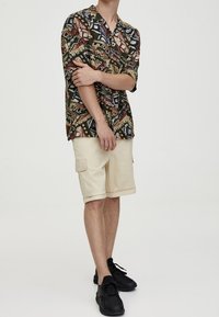 Camisa de mangas cortas con un patrón geométrico multicolor, pantalones cortos de cargo beiges y zapatillas negras. La camisa tiene un cuello y un corte relajado.