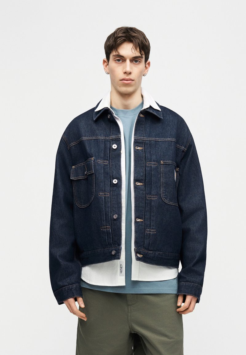 Junger Mann, der eine dunkelblaue Jeansjacke über einem weißen Hemd und einem hellblauen T-Shirt trägt, steht vor einem schlichten, hellgrauen Hintergrund.