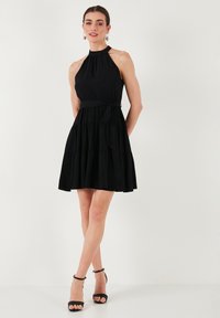 Robe noire sans manches avec un col haut, un corsage ajusté, une taille à nouer et une jupe à volants. Tissu lisse avec un design fluide. Sandales à talons noires.