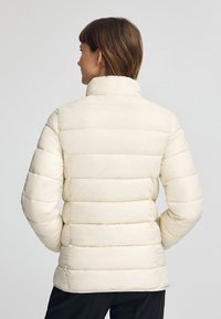 Vadderad jacka i ljus elfenben, med ståkrage, horisontell söm och långa ärmar; slät textur och figurnära silhuett.