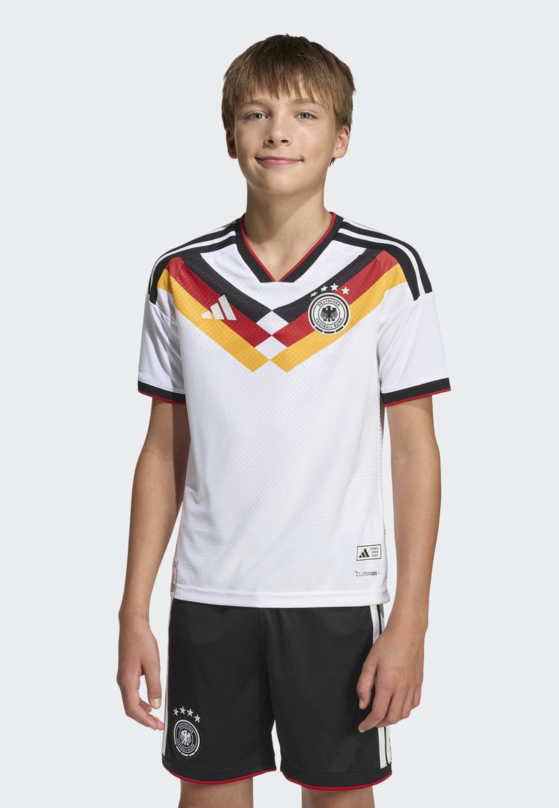 White soccer jersey with black, red, and yellow accents; v-neck design; geometric pattern; black shorts with logo and white stripes.