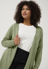 Cardigan lung, deschis în față, de culoare verde deschis, realizat dintr-un material moale. Purtați peste un tricou alb cu model în arabesc, asortat cu pantaloni negri. Are o croială lejeră.