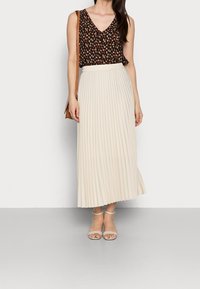 Top sin mangas negro con patrón floral, combinado con una falda midi plisada color crema y tacones transparentes de tiras. Bolso marrón usado en modo bandolera.