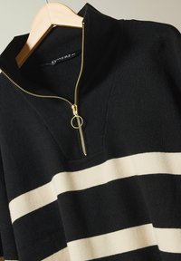 Schwarzer und cremefarbener gestreifter Strickpullover mit einem Reißverschlusskragen und goldenen Reißverschlussdetails. Der Stoff hat eine glatte Textur.