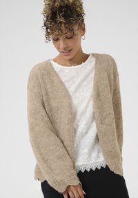 Beige Strickjacke mit lockerer Passform, hergestellt aus weichem Material. Sie hat überschnittene Schultern und einen leicht verkürzten Saum. Getragen über einer weißen Bluse mit Blumenmuster.