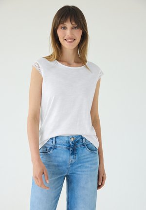 Kvinde smiler, iført hvid top med korte ærmer med blondekant og lyseblå jeans, står foran en ensfarvet hvid baggrund.