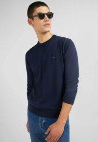 Pull en maille bleu marine à col rond avec des poignets côtelés, portant un petit logo sur la poitrine, associé à un jean en denim bleu clair.
