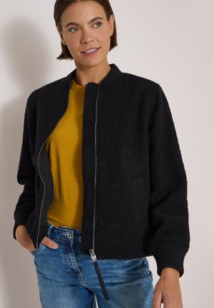 Femme portant une veste zippée noire texturée sur une chemise jaune moutarde avec un jean bleu, mains partiellement dans les poches, regardant légèrement sur le côté.