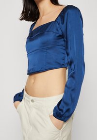 Krátká saténová blůza v tmavě modré barvě se čtvercovým výstřihem, volánkovými dlouhými rukávy a přiléhavým střihem. Měkká textura a čisté švy.