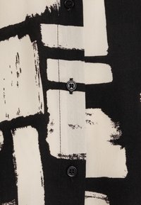 Materiał w czarno-białe wzory z abstrakcyjnym wzorem pędzla. Posiada czarny guzik na beżowym tle.