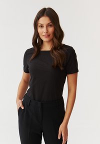 T-shirt noir à manches courtes avec un col carré, présentant une texture lisse, complété par un pantalon noir taille haute.