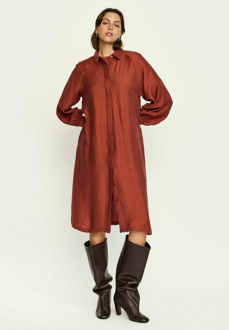 Lange, roestkleurige shirtdress van een getextureerde stof, met knoopsluiting en oversized mouwen, gecombineerd met kniehoge zwarte leren laarzen.