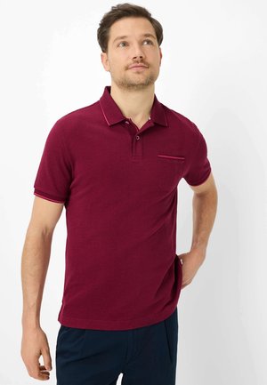 Mand iført en bordeaux kortærmet polo med en lomme, der kigger let opad med en hånd hvilende på hoften.