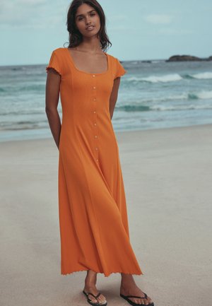 Nő áll a strandon, hosszú, gombos narancssárga maxiruha és fekete flip-flop papucsban, mögötte az óceán hullámai.