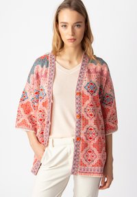 Daugiaspalvis lengvas kimono tipo švarkas su sudėtingais gėlių ir geometriniais raštais, turintis ryškų oranžinį sagų užsegimą ir plačias rankoves.
