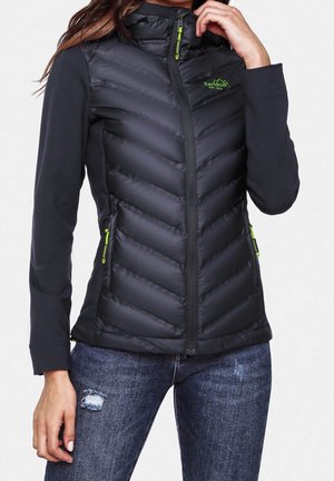 Femme portant une veste matelassée noire avec des tirettes de fermeture éclair néon vertes et un logo, assortie à un jean bleu déchiré.