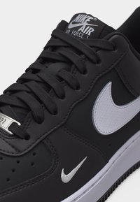 Μαύρο αθλητικό παπούτσι Nike Air Force 1 με λευκά λογότυπα Nike Swoosh στο πλάι και τη μύτη, μαύρα κορδόνια και ετικέτα με το brand στη γλώσσα.