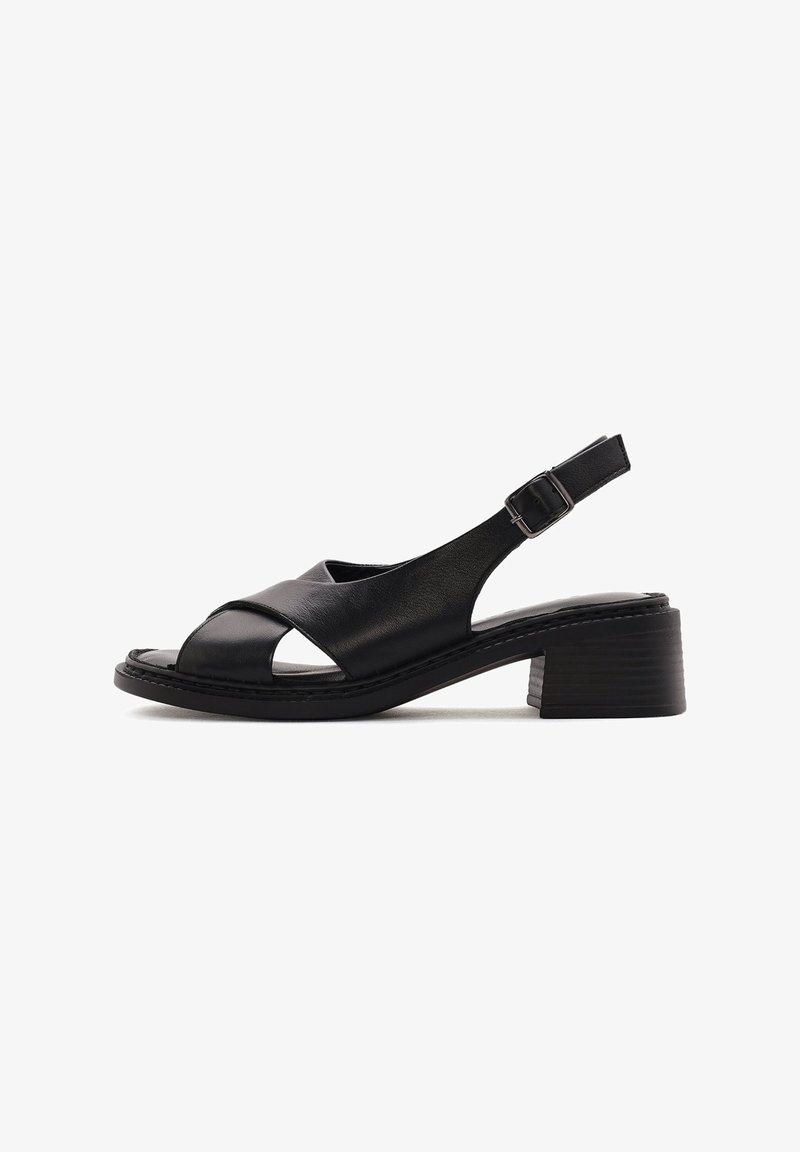 Sandale noire en cuir à bout ouvert avec brides croisées, boucle de cheville réglable et petit talon bloc sur fond blanc.