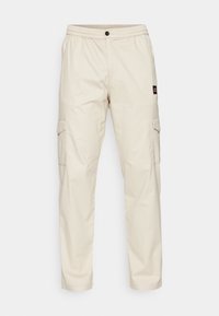 Pantalon cargo beige avec une ceinture élastique, une fermeture à bouton et deux poches latérales. Comprend une petite étiquette de marque sur la cuisse gauche.