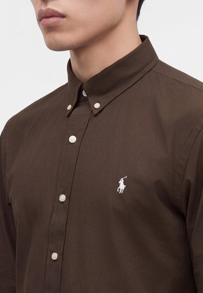 Homme portant une chemise marron boutonnée avec un col boutonné et un logo joueur de polo brodé blanc sur la poitrine.