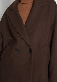 Brun dobbeltbrystet frakke med store sorte knapper, klapkrave og strukturerede skuldre, lavet af blødt stof. Ribbede brune turtleneck under.