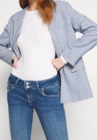 Blå och vit rutig kavaj över en vit ribbad topp, tillsammans med blå denimjeans med dubbelknäppning och lätt slitage.