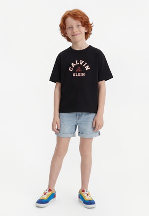 Κόκκινο-μαλλιαρό παιδί στέκεται, φοράει μαύρο T-shirt Calvin Klein, ανοιχτόχρωμα τζιν σορτς με ρεβέρ και πολύχρωμα αθλητικά παπούτσια, μπροστά σε απλό λευκό φόντο.