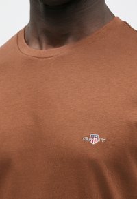 Brun bomulds-T-shirt med rund halsudskæring og korte ærmer. Bemærkelsesværdigt broderet GANT-logo på brystet. Blød tekstur med en almindelig pasform.