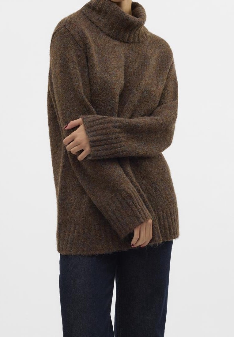 Personne portant un pull à col roulé marron oversize avec poignets et ourlet côtelés, associé à un pantalon bleu foncé sur fond uni.