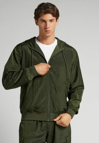 Chaqueta liviana verde oliva con cremallera, acabado texturizado, capucha y puños elásticos, combinada con una camiseta blanca debajo.