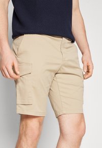 Homme portant un short cargo beige et une chemise texturée navy, les mains détendues sur les côtés, coupé à mi-thigh, fond neutre.