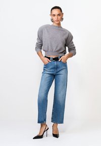 Femeie îmbrăcată cu un pulover gri scurt, blugi bleumarin cu talie înaltă și curea neagră, purtând pantofi cu toc stiletto negri, stând în fața unui fundal alb.