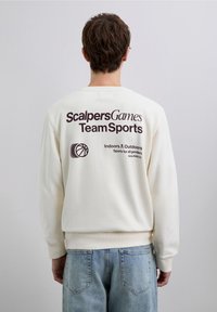 Krämfärgad sweatshirt med ribbade muddar och fåll. Bakstycket pryds av tryck med texten "Scalpers Games Team Sports" och en basketbollsgrafik.