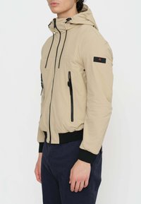 Giacca beige con cappuccio, zip e dettagli neri. Presenta un design aderente, polsini elasticizzati e una vita texturizzata.