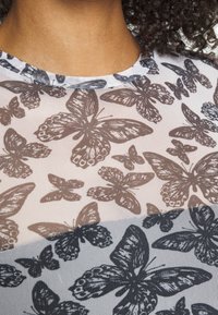 Närbild av person som bär en genomskinlig topp med detaljerade bruna och svarta fjärilar, lockigt hår synligt vid nacklinjen.