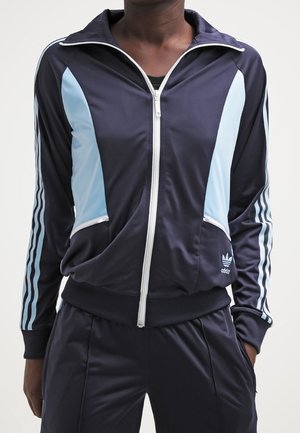 Person trägt einen dunkelblauen Adidas-Trainingsanzug mit hellblauen Einsätzen und weißen Streifen an den Ärmeln, steht mit den Händen in den Taschen.