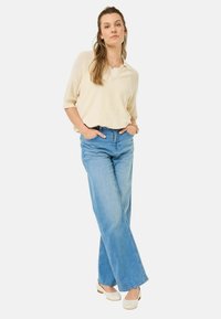 Pull v-neck beige clair associé à un jean large bleu clair taille haute. Le mannequin se tient avec les mains dans les poches, portant des sabots crème.
