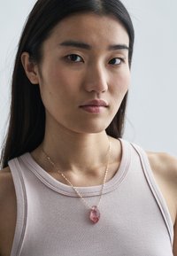 Colar dourado com um pendente de cristal bruto rosa, usado sobre um top rosa claro sem mangas, destacando a forma orgânica e a cor do colar.