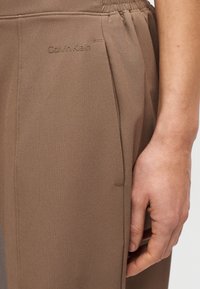 Καφέ παντελόνι από ύφασμα Calvin Klein με λεία υφή, διαθέτει πλαϊνές τσέπες, σούρα στη μέση και διακριτικό λογότυπο στο μπροστινό μέρος.