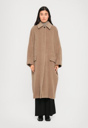 Woman with long dark hair wearing a loose, knee-length beige coat over black pants and black shoes, standing against a plain background.