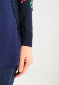 Marineblauer Cardigan mit floraler Stickerei auf dem Ärmel, aus einem weichen Strickmaterial gefertigt, mit lässiger Passform und glatter, eleganter Textur.